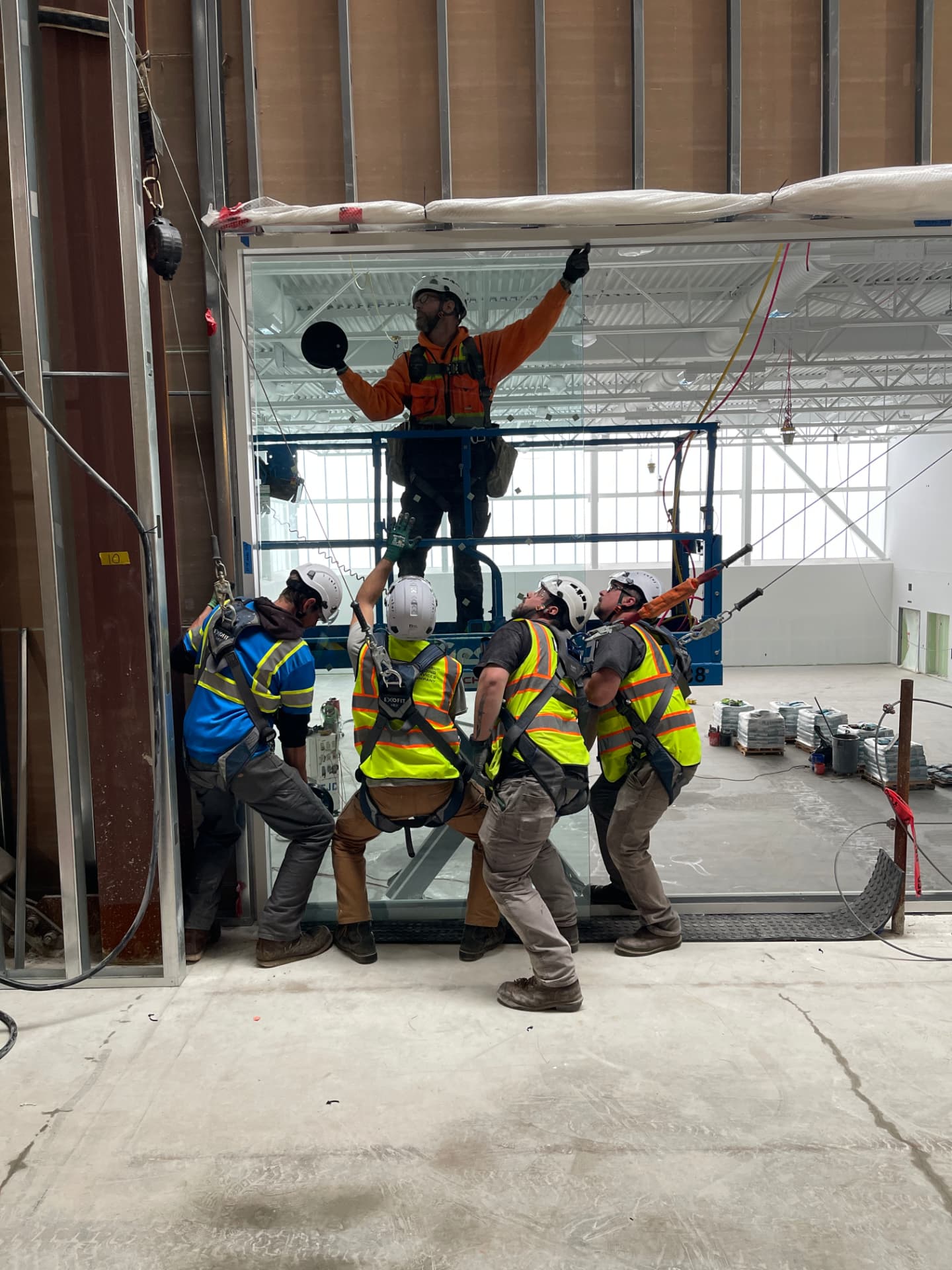 Crew of glaziers setting a large glass partition panel into its overhead track, one worker elevated on a scissor lift